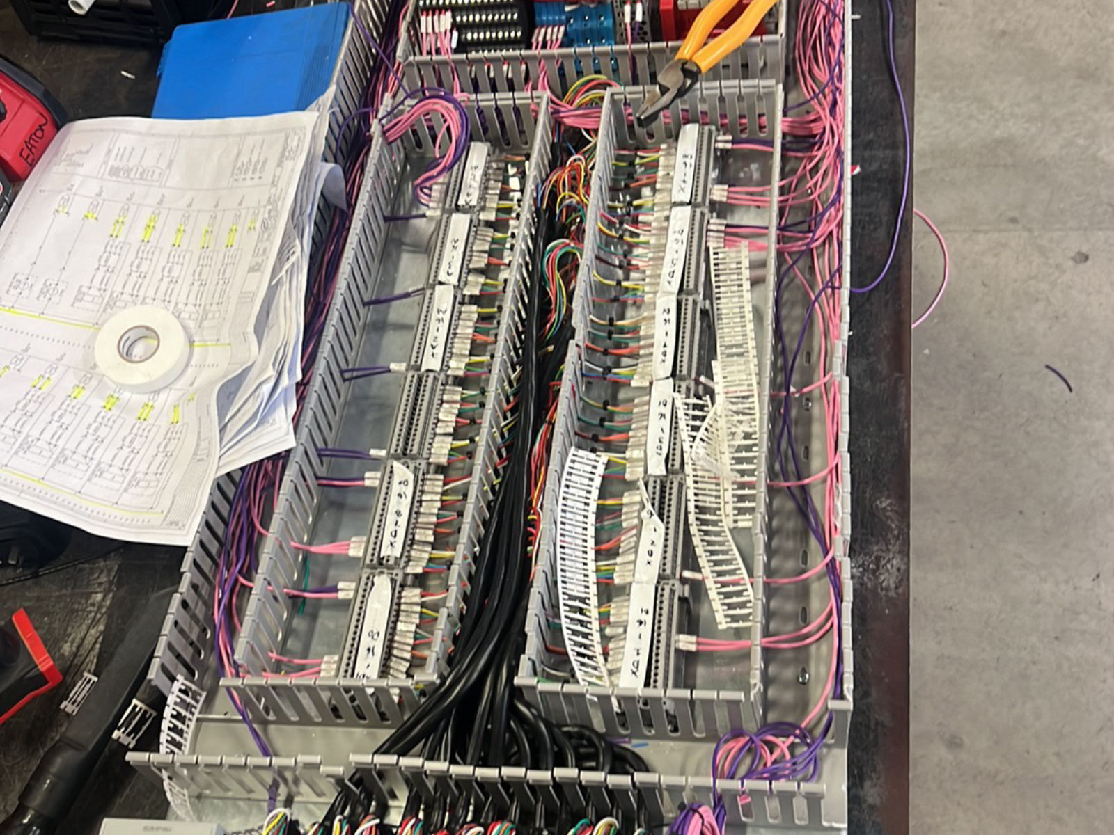 Switchboard gear plate on the bench with terminal rails, ferruled cores and wiring drawings alongside